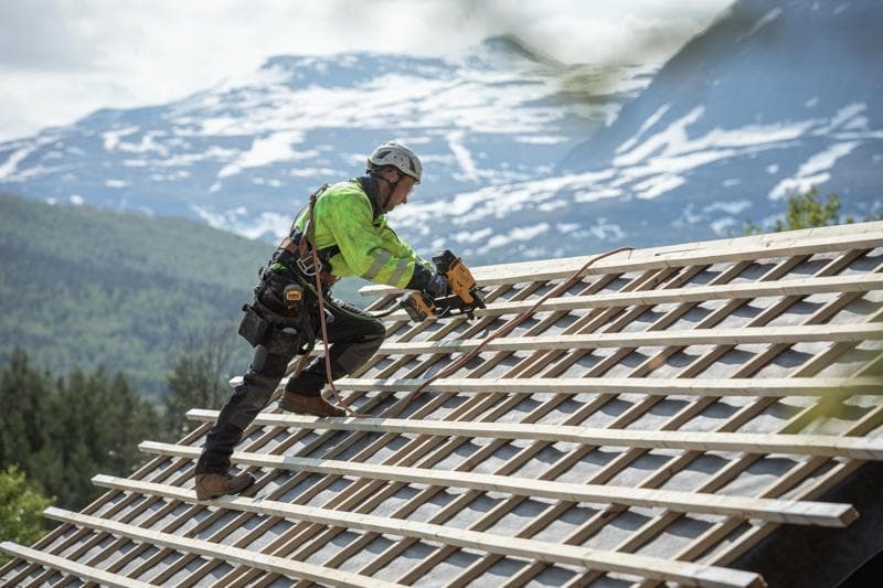Arbeiter nutzt einen DEWALT Nagler auf einem schrägen Dach im Freien, mit Sicherheitsausrüstung und Bergen hinten.