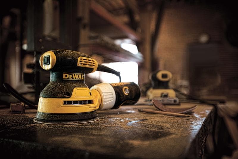 Nahaufnahme einer gelb-schwarzen DEWALT Elektroschleifmaschine auf einer staubigen Werkbank in einer Werkstatt. Im Hintergrund sind eine weitere Schleifmaschine und Schleifblätter unscharf zu sehen. Die Umgebung ist industriell mit Regalen und Werkzeugen.