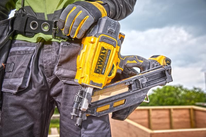 Person mit DEWALT DCN930N Nagelpistole, Handschuhen und Arbeitskleidung an einer Baustelle im Freien.