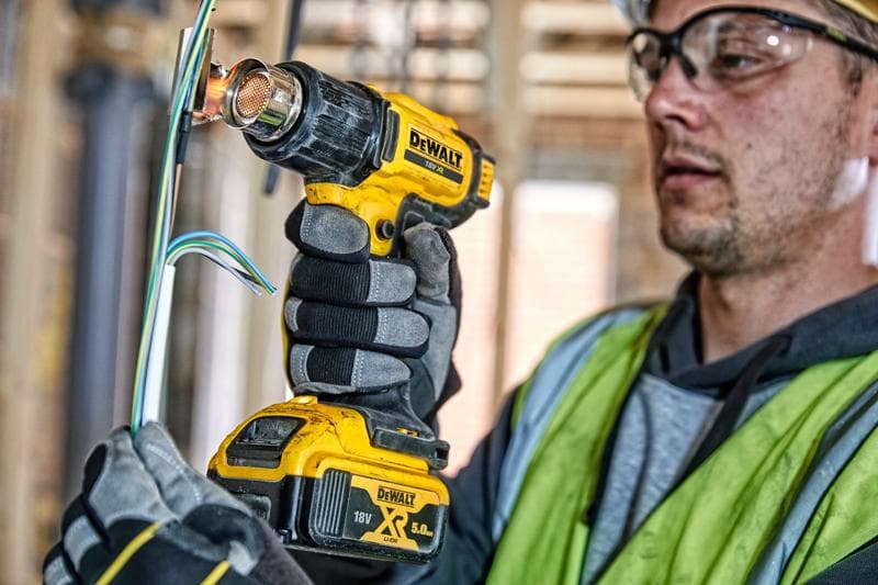 Eine Person mit Arbeitshandschuhen und Sicherheitsweste verwendet eine DEWALT DCE530N kabellose Heißluftpistole, um elektrische Kabel auf einer Baustelle zu erhitzen. Das Gesicht der Person ist aus Datenschutzgründen verschwommen.