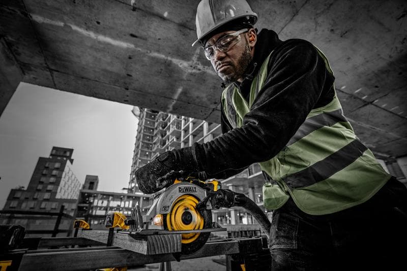 Person nutzt DEWALT DCS565NT Kreissäge auf Baustelle, trägt Schutzkleidung und sägt Holz.