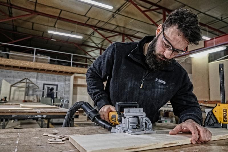 Eine Person verwendet ein DEWALT DW682K Holzwerkzeug, um ein Stück Holz auf einem Tisch in einer Werkstatt zu schneiden. Das Gesicht der Person ist aus Datenschutzgründen unkenntlich gemacht.