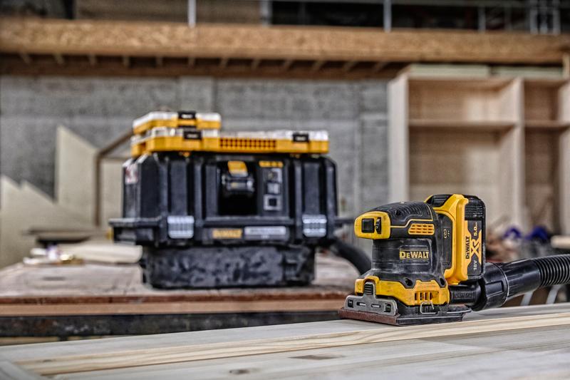 DEWALT Akku-Schleifer auf Holz, mit Aufbewahrungsbox im Hintergrund in einer Werkstatt.