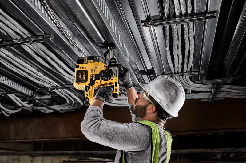 Eine Person mit Schutzhelm, Schutzbrille, Handschuhen und Sicherheitsweste benutzt einen gelben kabellosen DEWALT-Bohrhammer, um eine Metalldecke mit elektrischer Verkabelung zu bohren.