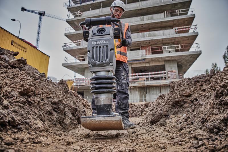 DEWALT Verdichter im Einsatz auf Baustelle, bedient von Arbeiter mit Schutzkleidung, umgeben von Erde und Gebäuden.